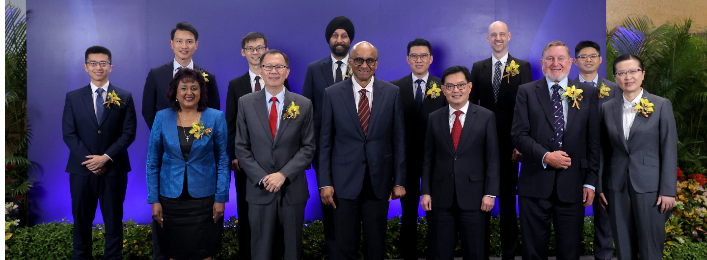 Group of thirteen people in formal attire with corsages, posed against a blue backdrop and foliage.